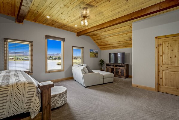 A spacious living room with a high ceiling, large windows with mountain views, a white sofa, a wooden TV stand, and a dining table. - Bear Island: Beautiful Lake Front Home w/ hot tub! Pet Friendly! (Pagosa Springs)