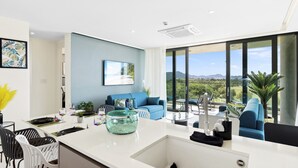 A modern kitchen with a large island, a dining area with chairs, a blue sofa, and a view of the outdoors through large windows.