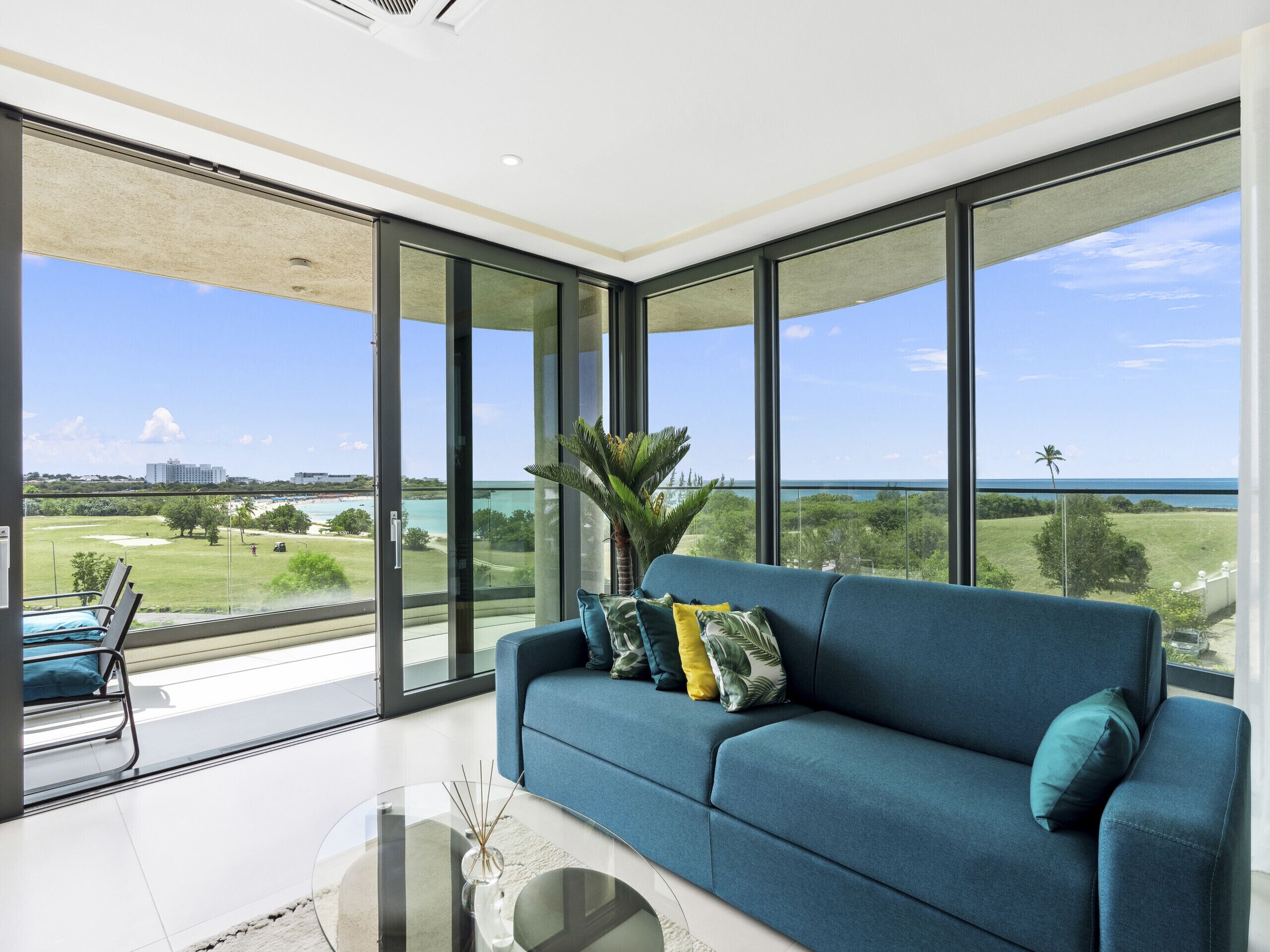 Un salon moderne avec un canapé bleu, une table basse en verre et de grandes fenêtres donnant sur la plage et l’océan.