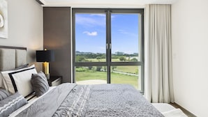 A modern bedroom with a large bed, a nightstand, a lamp, and a view of a green landscape through a sliding glass door.
