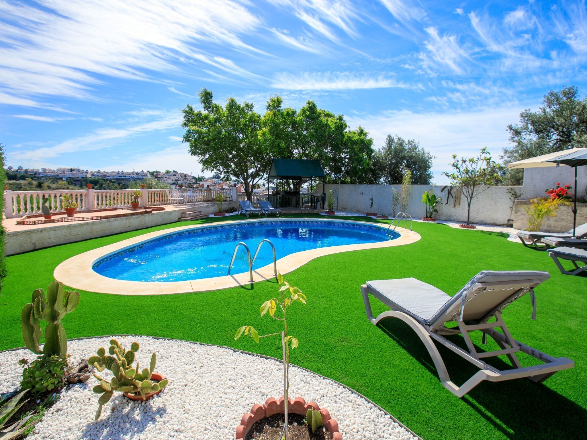 A swimming pool with a green lawn, a covered seating area, and a view of the city in the background.