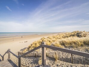 Ein Strand mit einer Holztreppe, die zum Sand führt, einer Sanddüne und dem Meer im Hintergrund.