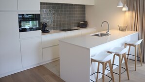 A modern kitchen with white cabinetry, a black backsplash, a built-in oven, a sink, and a bar area with white stools.