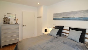 A bedroom with a bed, a nightstand, a lamp, a painting on the wall, and a dresser with a lamp and decorative items. - Holiday Home Backbert Near Kappeln (Kappeln)