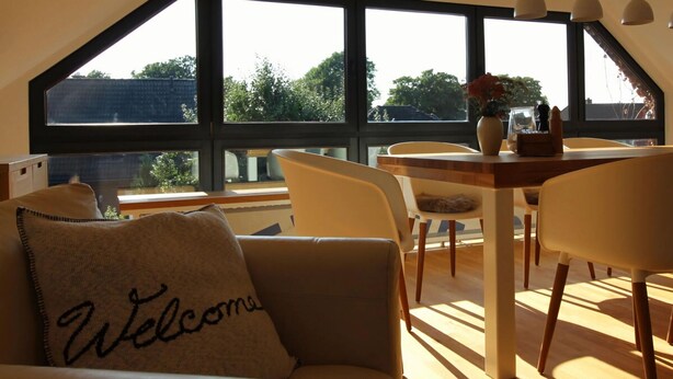 A modern living room with a sloped ceiling, large windows, a sofa with a 'Welcome' pillow, and a dining area with chairs and a table. - Holiday Home Backbert Near Kappeln (Kappeln)