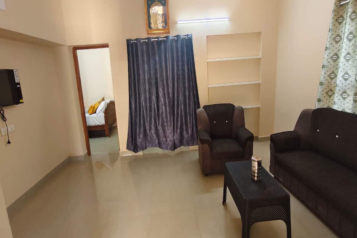 A living room with a sofa, armchair, and a small table. There is a TV mounted on the wall and a curtain.