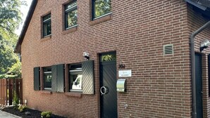 A two-story brick building with a black door, a mailbox, and a small garden with shrubs. - Florentine - Marbi (Varel)