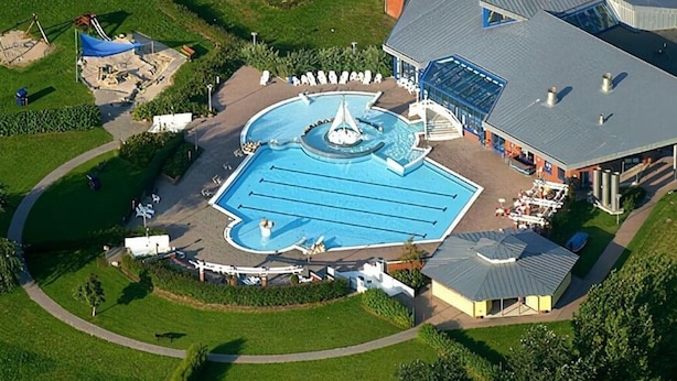 An aerial view of a swimming pool complex with a central fountain, surrounding lounge areas, and adjacent buildings. - Florentine - Marbi (Varel)