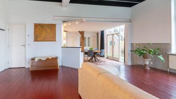 Een ruime woonkamer met houten vloer, een bank, een eettafel met stoelen en een groot raam dat zorgt voor natuurlijk licht.