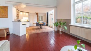 A spacious room with a wooden floor, a dining area with a table and chairs, a kitchen with white cabinets, and a large window with a view of trees. - Comfortable Farmhouse With Terrace (Nijeveen)