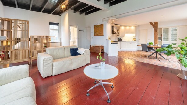 A spacious living area with a white sofa, a round coffee table, and a wooden floor. There is a kitchen area with white cabinets and a dining table. - Comfortable Farmhouse With Terrace (Nijeveen)