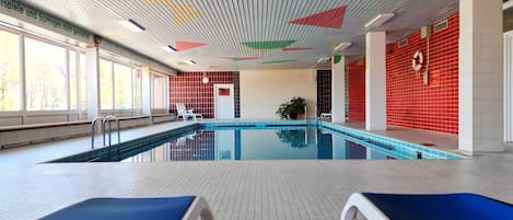 An indoor swimming pool with blue lounge chairs, a tiled floor, and a ceiling with colorful geometric patterns.