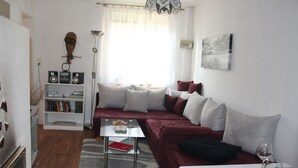 Una sala de estar con un sofá, una mesa de centro y una mesa de comedor servida con platos y copas de vino. Hay una estantería con libros y una lámpara.