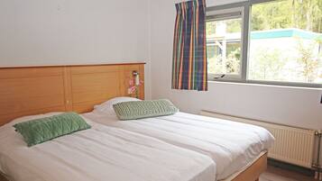 A room with two beds, a window with curtains, a radiator, and a view of greenery outside.