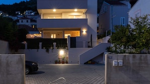 A modern two-story building with a flat roof, a balcony on the upper floor, and a gated entrance with a driveway leading to a parked car. - Apartments Amare - Three Bedroom Apartment With Terrace and Jacuzzi (Dubrovnik)