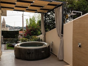 A hot tub in a covered outdoor area with a pergola overhead, a showerhead, and a view of a town in the background. - Apartments Amare - Three Bedroom Apartment With Terrace and Jacuzzi (Dubrovnik)
