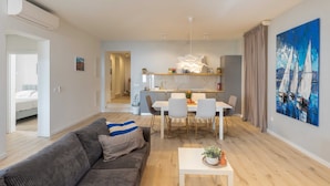 A modern living room with a grey sofa, a white coffee table, and a dining area with a white table and chairs. There is a kitchen in the background with a white countertop and a painting of sailboats on the wall. - Apartments Amare - Three Bedroom Apartment With Terrace and Jacuzzi (Dubrovnik)