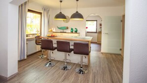 A modern kitchen with a central island, bar stools, pendant lighting, and a view of the garden through a window.