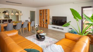 A modern living room with a large orange sofa, a coffee table, and a TV. There is a dining area with chairs and a table, and a bookshelf filled with books and decorative items.