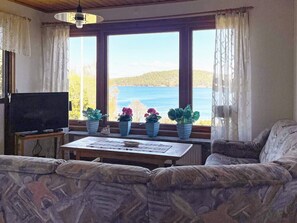 A living room with a sofa, a television, a table with papers, and potted plants by a window overlooking a body of water. - 5 Person Holiday Home in Stillingson (Stillingsön)