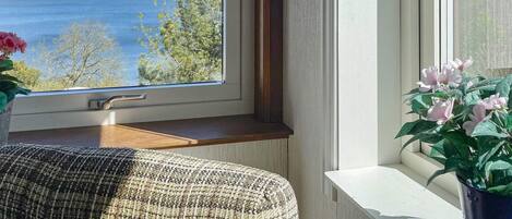A room with a view of a lake and hills, a window sill with a plant, and a piece of furniture.