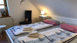 Een slaapkamer met een bed voorzien van een dekenovertrek met nautisch thema, een nachtkastje met een lamp, een stoel, en een klein tafeltje met een fles en een vaas.
