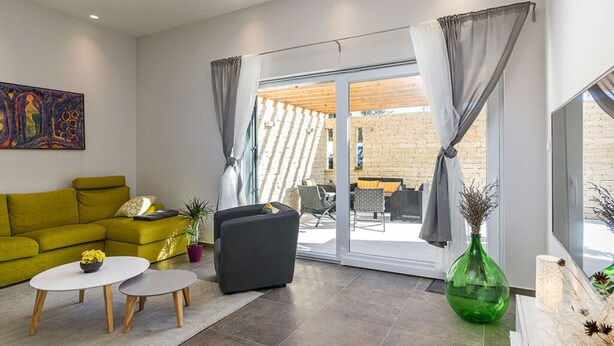 Een moderne woonkamer met een groene bank, een grijze fauteuil en een salontafel. Er is een schuifdeur die leidt naar een terras met een bakstenen muur en tuinmeubilair.