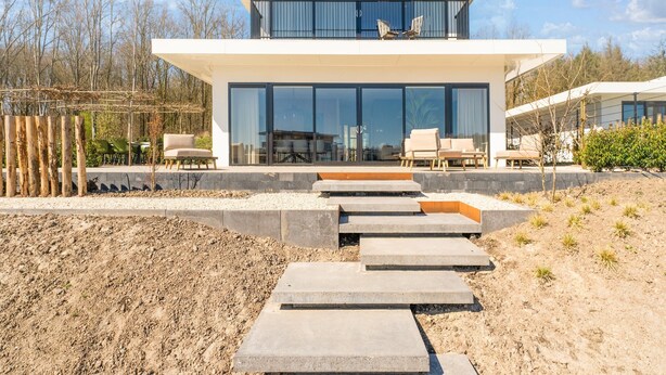 Een modern huis met twee verdiepingen, een balkon, terrasmeubilair en een pad van stenen dat naar de ingang leidt.