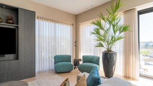 Een moderne woonkamer met een grote flatscreen televisie, twee blauwgroene fauteuils, een grote potplant en een bank met blauwe en beige kussens.