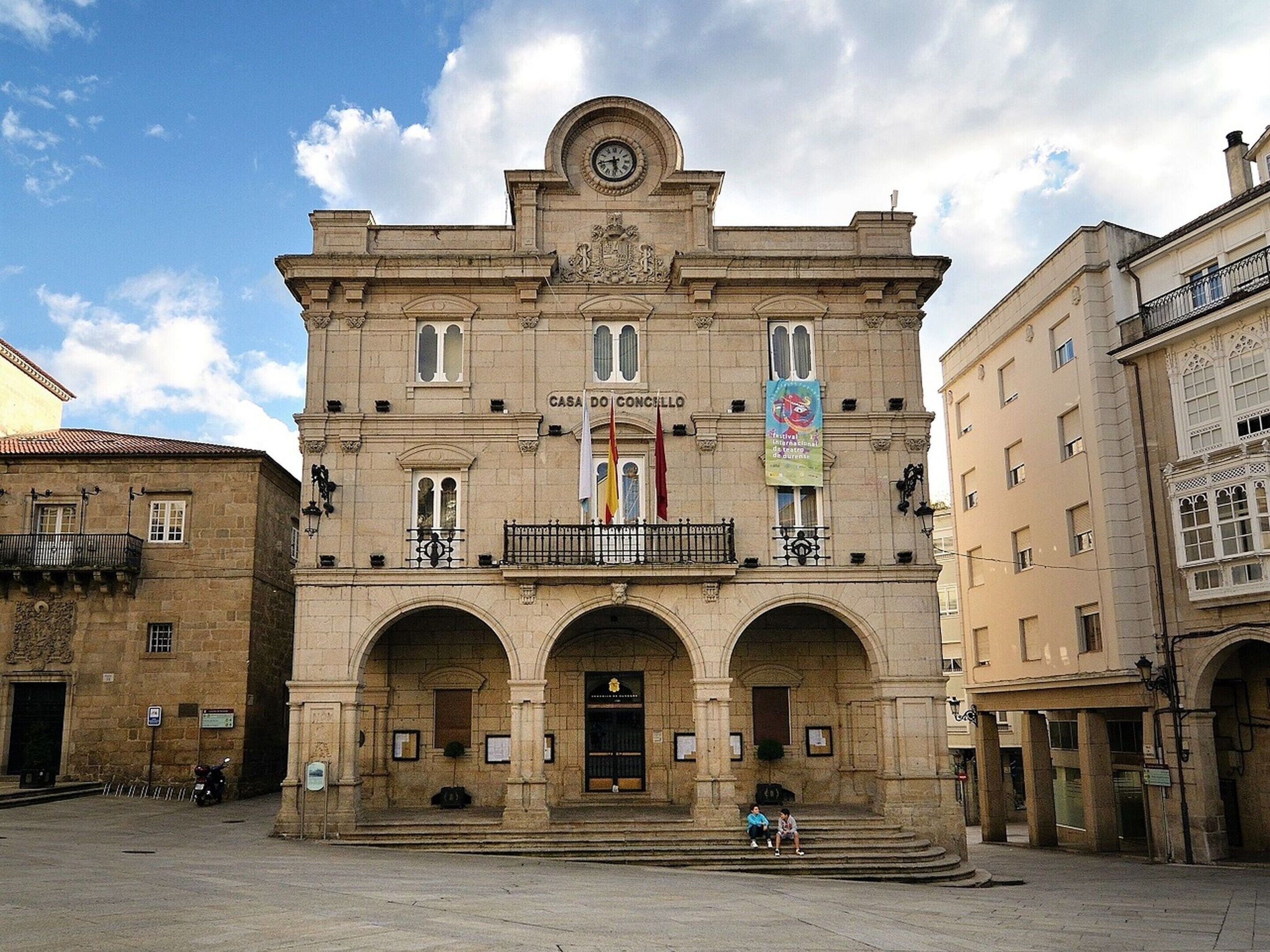 Un bâtiment historique doté d’une tour horlogée, d’entrées en arc de cercle et de drapeaux.