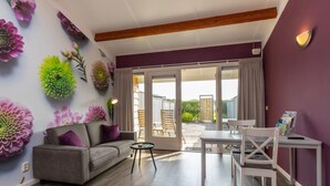 Un salon comprenant un canapé, une table et des chaises, une porte-fenêtre coulissante donnant sur un espace extérieur, et des fleurs décoratives sur le mur.