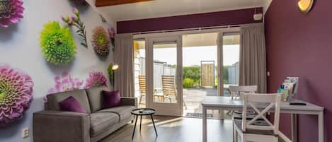 A living room with a sofa, a table, and chairs, a sliding glass door leading to an outdoor area, and decorative wall art.