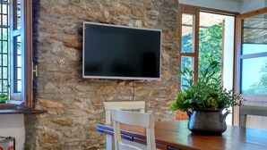 Une cuisine avec un mur en pierre, un téléviseur à écran plat fixé au-dessus d’un comptoir en bois, une chaise blanche et une plante en pot.