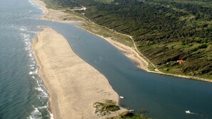 Vue aérienne d’une zone côtière avec une plage de sable, un étroit chenal et, au loin, des collines densément boisées.