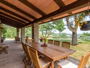 A covered patio with a wooden table and chairs, surrounded by brick flooring and potted plants.