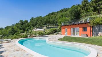A swimming pool with a stone deck, a small building with a red exterior, and a grassy area with trees in the background.
