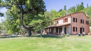 Una casa de dos pisos con fachada de ladrillo y techo de tejas, rodeada de un césped cuidado y árboles grandes.