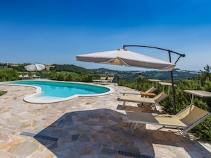 A swimming pool with stone tiles, surrounded by lounge chairs and umbrellas, with a view of green hills in the background.