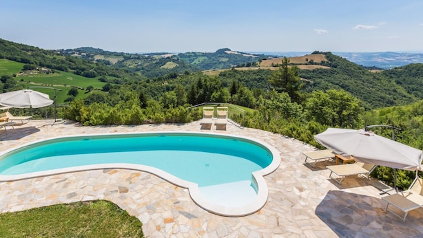 Una piscina con cubierta de piedra, rodeada de sillas de descanso y sombrillas, con vistas a un paisaje montañoso.