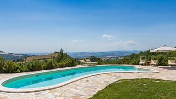 A curved swimming pool with stone tiles, surrounded by a grassy lawn and trees, with a view of distant hills and a clear sky.