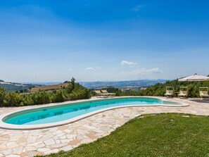 Une piscine aux bords incurvés, revêtue de carreaux de pierre, entourée d’une pelouse verdoyante et d’arbres, offrant une vue sur des collines lointaines et un ciel dégagé.
