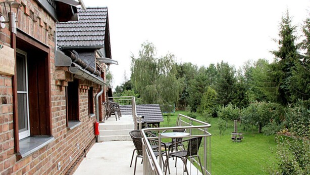 A brick building with a covered outdoor seating area, including tables and chairs, and a grassy yard with trees. - Pear - On the Fruit Tree (Golssen)