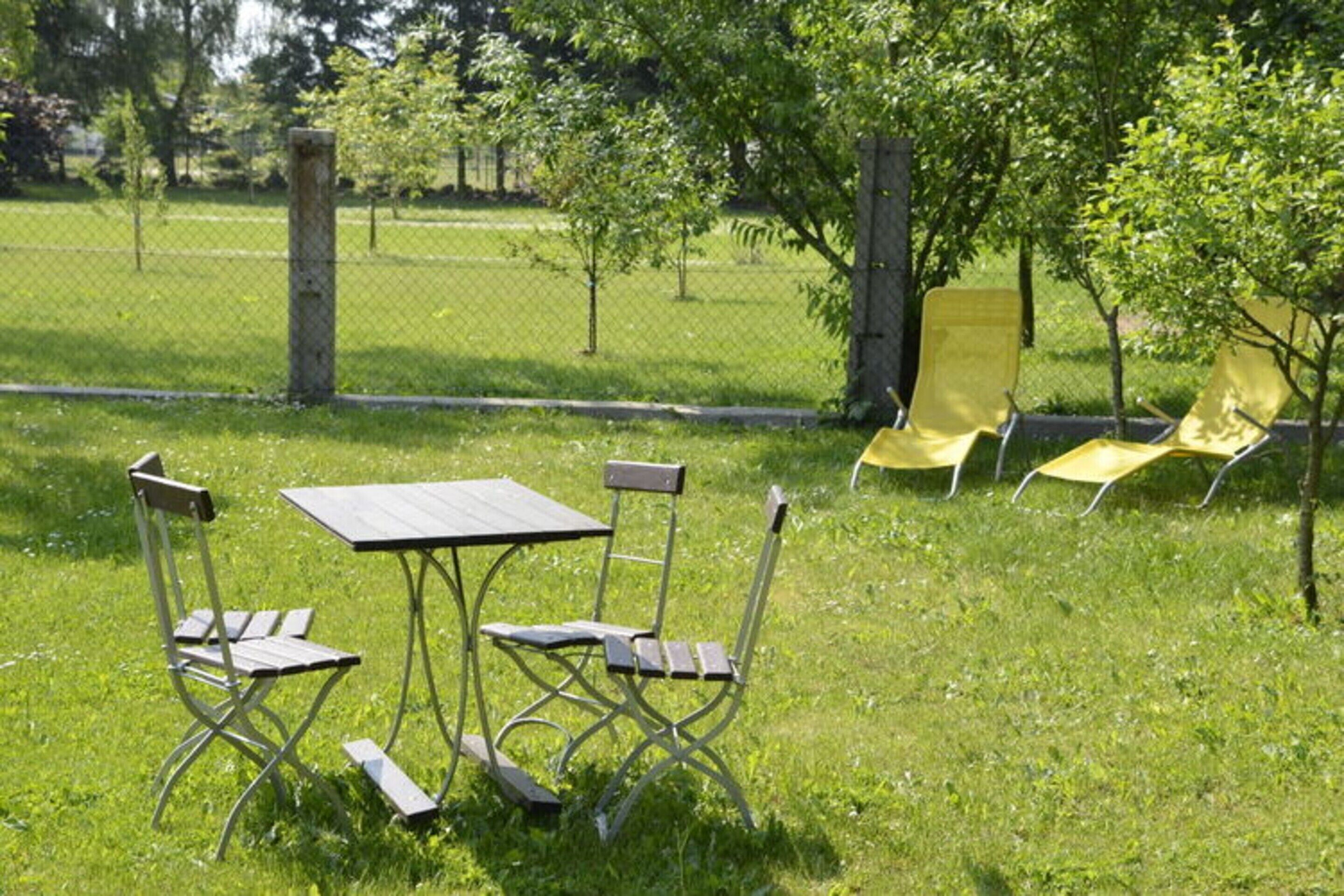 Ein sonniger Außenbereich mit Rasen, einem Metalltisch und vier Stühlen sowie zwei gelben Liegestühlen.