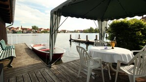 Une terrasse en bois avec une table et des chaises, un petit bateau et une vue sur une rivière, avec des bâtiments en arrière-plan.