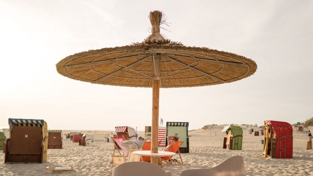 A beach with a thatched umbrella, beach chairs, and colorful beach huts. - Moi"n Dag Modern Retreat (Borkum)