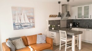A modern kitchen with white cabinets, a dining area with a white table and chairs, and a living area with a brown sofa and a small side table. - Moi"n Dag Modern Retreat (Borkum)