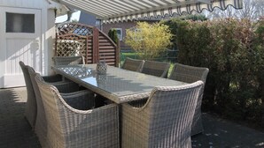 Un espace extérieur aménagé pour les repas, avec des meubles en osier et une table en verre, entouré d’un bâtiment blanc et d’une clôture en bois.