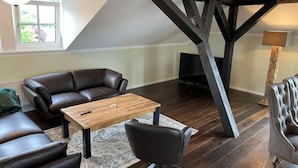 Un salon chaleureux avec un canapé en cuir marron foncé, une table basse en bois et un fauteuil noir. Une grande fenêtre laisse entrer la lumière naturelle, et un téléviseur est fixé au mur.