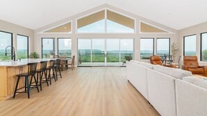 Een ruime woonkamer met een groot raam met uitzicht op een prachtig landschap, een keuken met barstoelen, en een zithoek met lederen fauteuils.