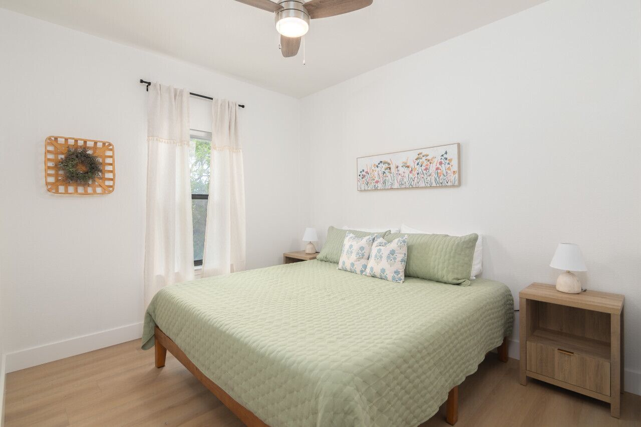 Een slaapkamer met een bed, nachtkastje, plafondventilator, raam met gordijnen en een wanddecoratie.